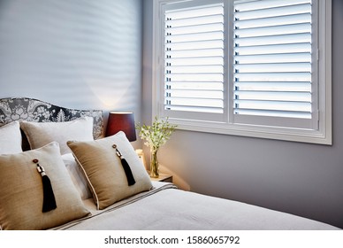Interior photography of neutral toned bedroom with white bedding, upholstered bed head, tasseled cushions, bedside table and lamp, flowers and window with plantation shutters