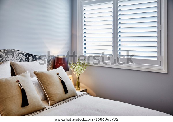 Interior\
photography of neutral toned bedroom with white bedding,\
upholstered bed head, tasseled cushions, bedside table and lamp,\
flowers and window with plantation\
shutters