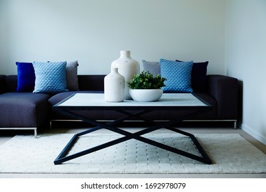 Interior Photography Of A Simple Modern Lounge Room With A Dark Blue Modular Sofa With Cushions, A Stone Topped Coffee Table With Decorative Bottles & A Succulent Plant, Shot Front On