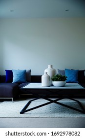 Interior Photography Of A Simple Modern Lounge Room With A Dark Blue Modular Sofa With Cushions, A Stone Topped Coffee Table With Decorative Bottles & A Succulent Plant, Shot Front On