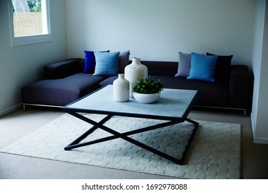 Interior Photography Of A Simple Modern Lounge Room With A Dark Blue Modular Sofa With Cushions, A Stone Topped Coffee Table With Decorative Bottles & A Succulent Plant, Shot Front On