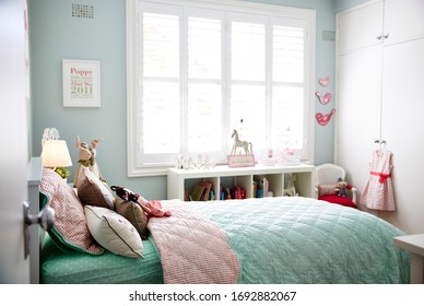 Interior Photography Of A Stylish Little Girl's Bedroom With Aqua And Pink Bedding, Decorative Cushions, Side Table With Lamp, Bookshelf, Wall Art And Toys