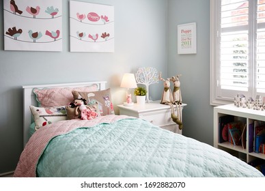 Interior Photography Of A Stylish Little Girl's Bedroom With Aqua And Pink Bedding, Decorative Cushions, Side Table With Lamp, Wall Art, Bookshelf And Toys