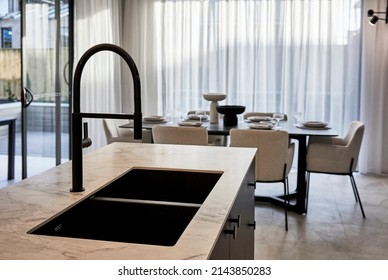 Interior Photography Of A Suburban Family Home, A Black Tap And Kitchen Sink In A Quartz Stone Bench Top Counter Top Within A Modern Kitchen, A Dining Room And Al Fresco Area In The Background.