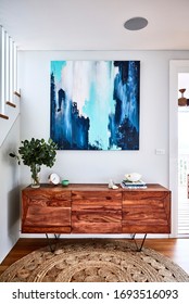 Interior Photography Of A Timber Sideboard With Decorative Objects, Gum Leaves In A Vase And An Abstract Painting With A Staircase On The Left And A Jute Rug On The Floor