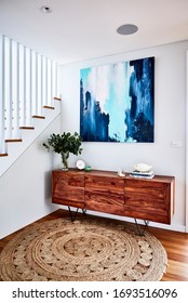 Interior Photography Of A Timber Sideboard With Decorative Objects, Gum Leaves In A Vase And An Abstract Painting With A Staircase On The Left And A Jute Rug On The Floor