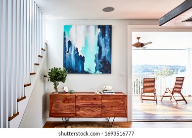 Interior Photography Of A Timber Sideboard With Decorative Objects, Gum Leaves In A Vase And An Abstract Painting With A Staircase On The Left And A Jute Rug On The Floor, A Timber Deck On The Right