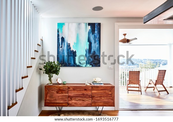 Interior photography of a timber sideboard with
decorative objects, gum leaves in a vase and an abstract painting
with a staircase on the left and a jute rug on the floor, a timber
deck on the right