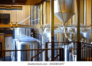 The interior production room of a microbrewery. Multiple large stainless steel brewing vats with metal pipes, hoses and fermentation tanks for handcrafted craft beer. The industrial facility is empty.