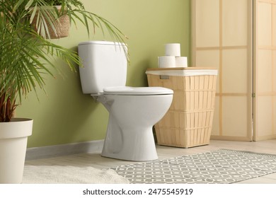Interior of restroom with toilet bowl, bin, folding screen and plant near green wall