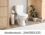 Interior of restroom with toilet bowl, plant and paper holder near grey wall