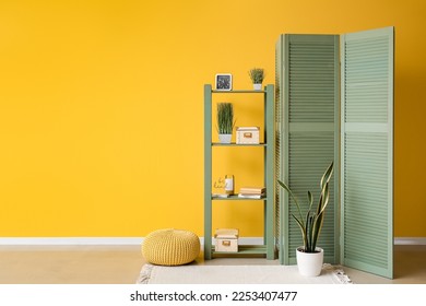 Interior of room with folding screen, shelving unit and houseplant near yellow wall