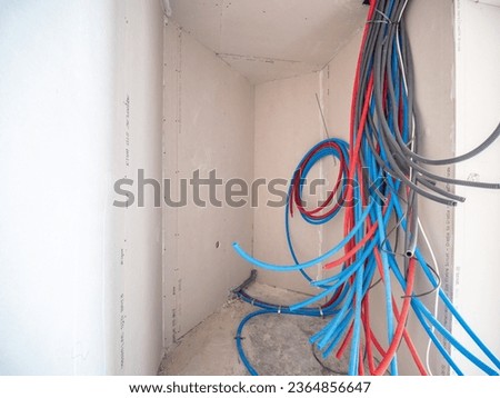 interior of a room, under construction, with red and blue plastic sheaths protruding from the wall and containing electrical cables 