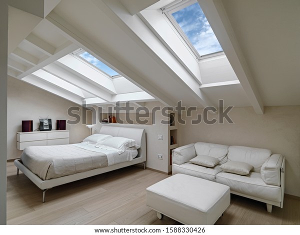 interior shot of a bedroom\
in the modern attic-room in the foreground the leather sofas with\
footrest in the background there is the bed illuminated by two\
skylights