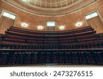 An interior shot of an old lecture hall with curved wooden seating and iron railings. The room features warm lighting, decorative walls, and a domed ceiling with windows