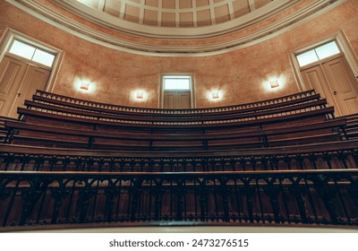 An interior shot of an old lecture hall with curved wooden seating and iron railings. The room features warm lighting, decorative walls, and a domed ceiling with windows - Powered by Shutterstock