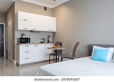 The interior of a small apartment with bed, blue pillows, light kitchen furniture, chairs with a table, kitchen appliances and refrigerator. apartment has gray walls.  fruits on the kitchen table. - Powered by Shutterstock