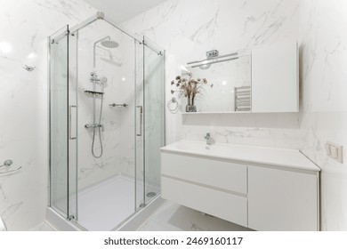 The interior of a small and minimalistic , white bathroom . A bedside table with a sink under the mirror and a shower cabin and white marble tiles .