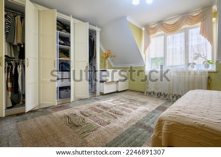 Interior of spacious bedroom in rustic style. Lot of light from a large window, double bed and large wardrobe closet with open doors showing inside