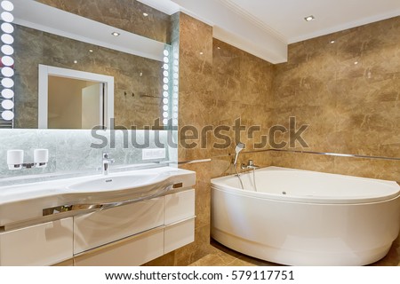 Interior of stylish bathroom in brown and white colors, with rounded ceramic bath in corner of the room and big mirror on wall and light around. White commode and accessories for bath.