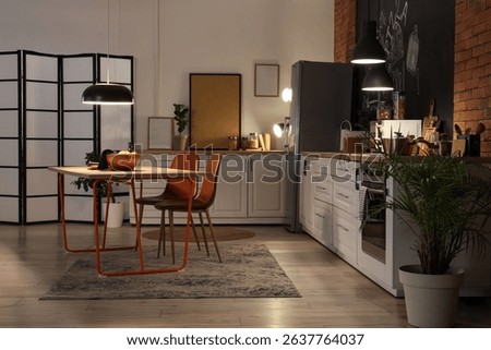 Interior of stylish kitchen with counters, dining table and glowing lamps at night
