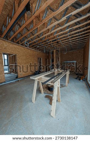 Interior of a timber frame under rough in shows engineered joists, OSB walls, conduits, sawhorse workbench, concrete slab, cables, scaffolding, and conduit cuts in natural light.