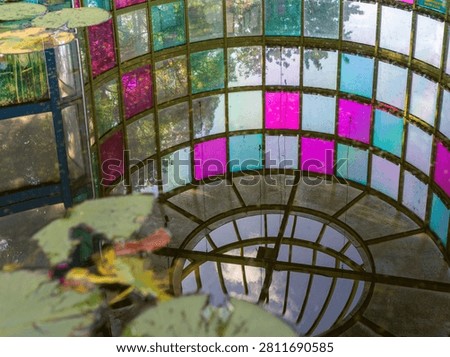 Interior upward perspective looking through a colorful grid stained glass dome structure with green lily pads overlaying the frame. The scene represents avant-garde architecture, vibrant color patterns, creative lighting, abstract concept, and artistic interior design.