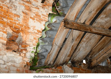 Interior view of an abandoned building featuring an old red brick wall and a collapsing roof made of wooden beams and corrugated sheets.