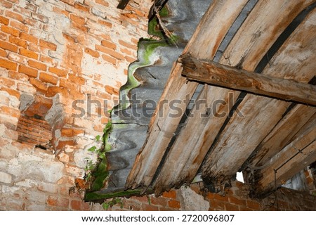 Interior view of an abandoned building featuring an old red brick wall and a collapsing roof made of wooden beams and corrugated sheets.