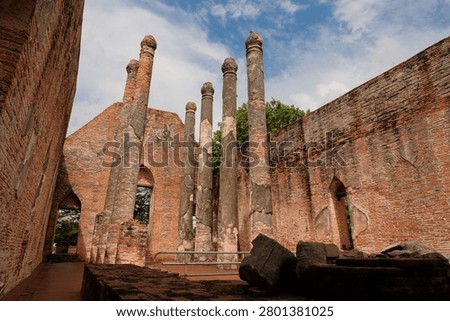 Interior view of the ancient assembly hall ruins at Wat Kudi Dao in Ayutthaya, Thailand. Tall weathered pillars stand among red brick walls and fallen debris under a beautiful bright cloudy blue sky