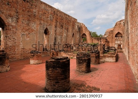 Interior view of the ancient assembly hall ruins at Wat Kudi Dao in Ayutthaya, Thailand. Red brick walls, arched windows, and short column bases sit on a tiled floor beneath a cloudy summer sky