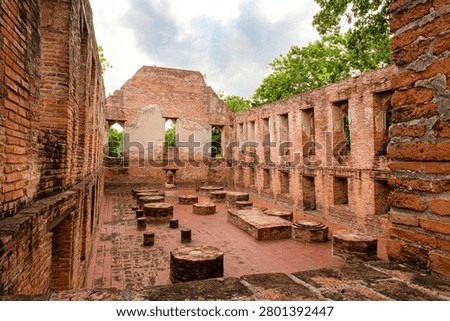 Interior view of the ancient assembly hall ruins at Wat Kudi Dao in Ayutthaya, Thailand. Red brick walls with rows of windows surround column bases on a tiled floor beneath a cloudy summer sky
