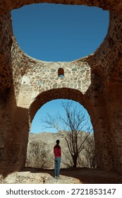 An interior view of the ancient Fire Temple of Natanz, Iran, framed by massive stone arches and an open circular roof. The structure is built from rough, irregular stones bonded with mortar, reflecting early Iranian masonry and pre-Islamic architectural traditions associated with Zoroastrian heritage. In the center of the composition, a single person is seen from behind, standing beneath the arch and looking outward toward a mountainous landscape and leafless trees, creating a strong sense of scale and contemplation. Natural sunlight pours through the open dome, illuminating the textured stone surfaces and producing dramatic contrasts between light and shadow. The clear blue sky above enhances the geometry of the arches and emphasizes the connection between architecture, nature, and spirituality. This image conveys themes of history, ancient religion, cultural heritage, solitude, and the enduring presence of Persian civilization.