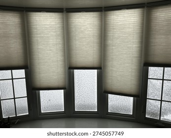 Interior view of bay windows with cellular shades partially lowered as heavy snow and frost cover the glass during a winter storm, creating a cold and cozy seasonal atmosphere inside a home.