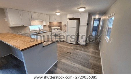 Interior view of a compact kitchen remodel featuring white shaker cabinets and a butcher block peninsula countertop. The space includes wood look laminate flooring and white appliances including a gas range and refrigerator. Main work surfaces utilize a grey stone pattern laminate countertop with a stainless steel sink. Simple flush mount ceiling lights illuminate the narrow functional layout typical of a small apartment or mobile home renovation.