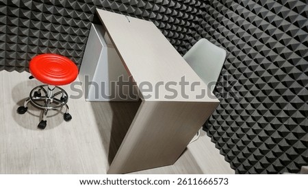 Interior view of a compact workspace. Includes a bright tabletop desk, a light gray chair, and a contrasting red adjustable stool. Acoustic treatment on the walls.