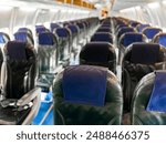 Interior view of empty airplane cabin with rows of black leather seats and blue headrest covers, illuminated by overhead lights, showing aisle running down middle and windows on both sides.