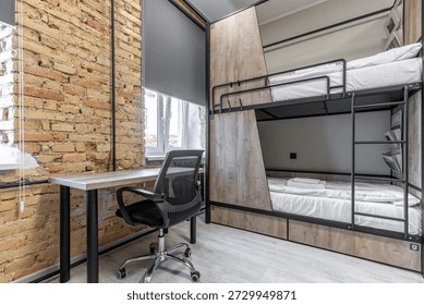 Interior view of a hostel dormitory featuring a black metal bunk bed with wood privacy panels and integrated storage. The room includes a wooden desk with a black mesh office chair and a rustic exposed brick accent wall.