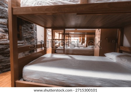 Interior view of hostel dormitory seen through wooden bunk beds, clean shared sleeping room with mattresses and pillows, budget travel accommodation, perspective depth, natural light, no people