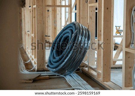 Interior view of a house under construction featuring wooden framing, exposed wiring, and a coiled ventilation duct on the floor under natural light.