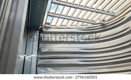Interior view of a metal basement window well with vertical steel ridges and a grated cover on top. Light filters through the grate, illuminating the steel walls and creating a sense of depth.