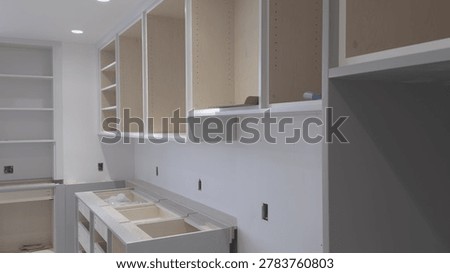 Interior view of a modern kitchen during cabinet installation. Unfinished light natural wood cabinet carcasses with gray painted frames and panels are shown in various stages of assembly. Features open shelving, drawer units, upper wall cabinets, and a countertop area. The space shows typical new construction or renovation details with visible electrical outlets.