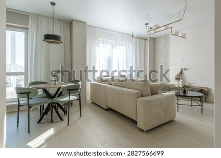 Interior view of a modern open-plan living room and dining area featuring a light beige sofa, a dining table with green upholstered chairs, large windows with sheer curtains, a projector screen on the wall, and stylish pendant lighting.