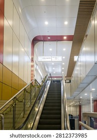 Interior view of a modern public facility featuring an empty escalator moving upwards alongside stairs. Brightly lit ceiling, architectural details, and overhead navigation signage emphasize a clean and contemporary transit or commercial space