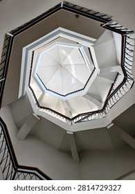 Interior view of a multi-story octagonal architectural structure featuring a central staircase, white walls, dark metal railings, and wooden handrails. The geometric design and skylight create a unique modern architectural composition.