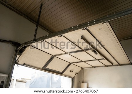 Interior view of an open sectional garage door with metal tracks and ceiling panels, showing workshop entrance and overhead mechanism.