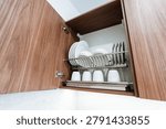 Interior view of an open walnut-finish upper kitchen cabinet fitted with a stainless steel two-tier dish rack holding stacked white plates and bowls on the