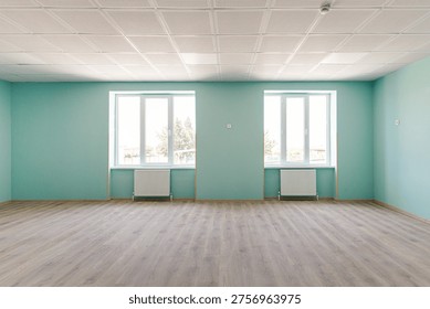 Interior view of a spacious empty hall featuring bright turquoise walls, two large windows with radiators, wooden laminate flooring and a professional acoustic grid ceiling.