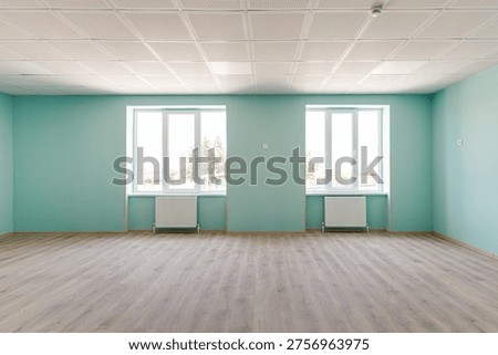 Interior view of a spacious empty hall featuring bright turquoise walls, two large windows with radiators, wooden laminate flooring and a professional acoustic grid ceiling.