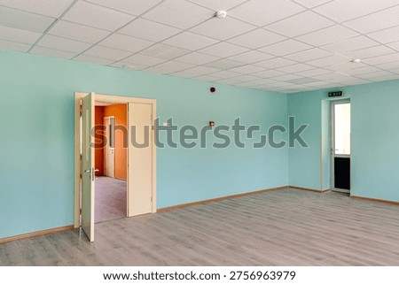 Interior view of a spacious empty hall featuring bright turquoise walls, two large windows with radiators, wooden laminate flooring and a professional acoustic grid ceiling.